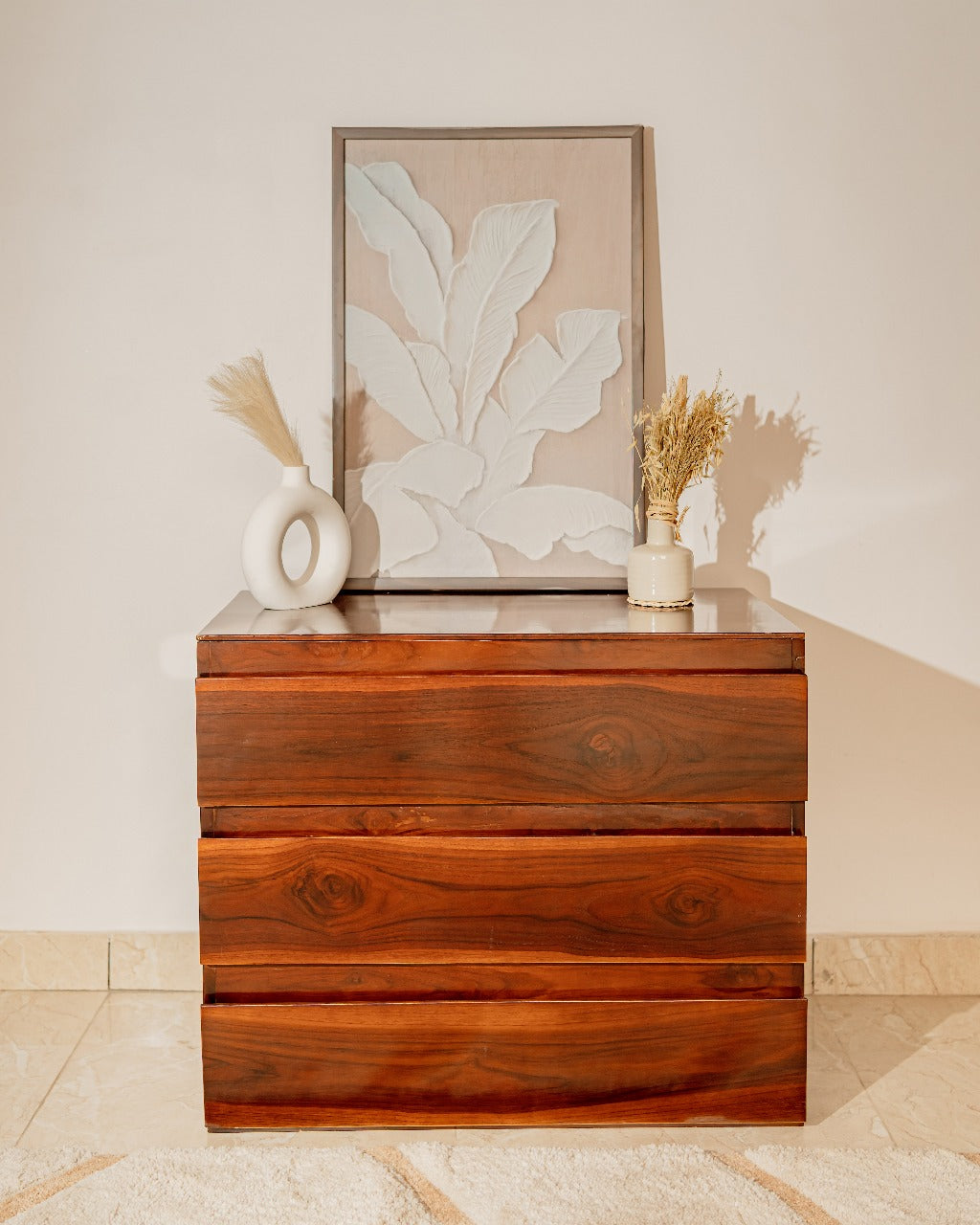 Wooden dresser with decorative items including a vase and artwork against a neutral background.